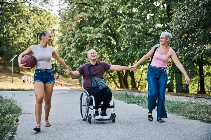 Man in wheelchair on a path outside holding hands with two female friends